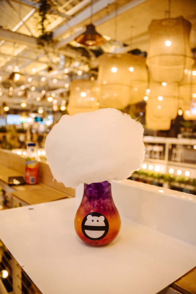 Candy floss served on top of an iced tea in a lightbulb-shaped glass, set on a white table with a warmly lit food court in the background