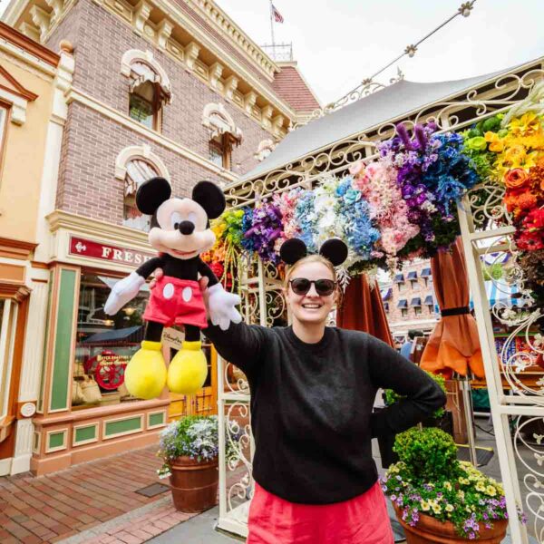 Person in sunglasses and a black top, posing with a plush Mickey Mouse in front of a colorful flower display at Disneyland California