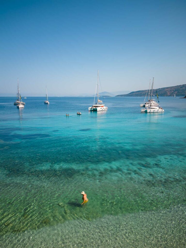 A drone captures the breathtaking clarity of the Ionian Sea at a Greek beach, with yachts anchored in the tranquil azure waters and a swimmer enjoying the idyllic surroundings