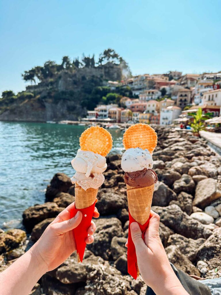 Two hands holding up delicious ice cream cones against the picturesque backdrop of a Greek beach, with sunlit waters and coastal village scenery