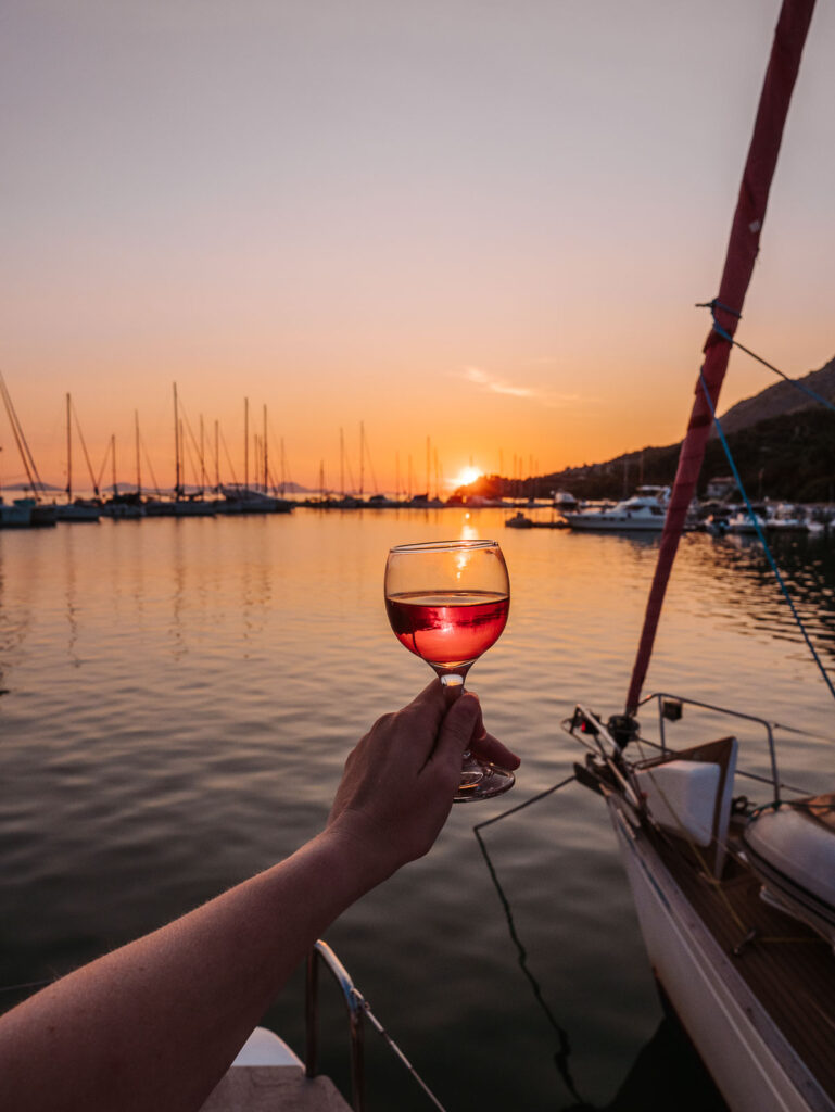 A hand holding a glass of wine against a beautiful sunset at a calm marina, with silhouettes of moored sailboats enhancing the tranquil scene