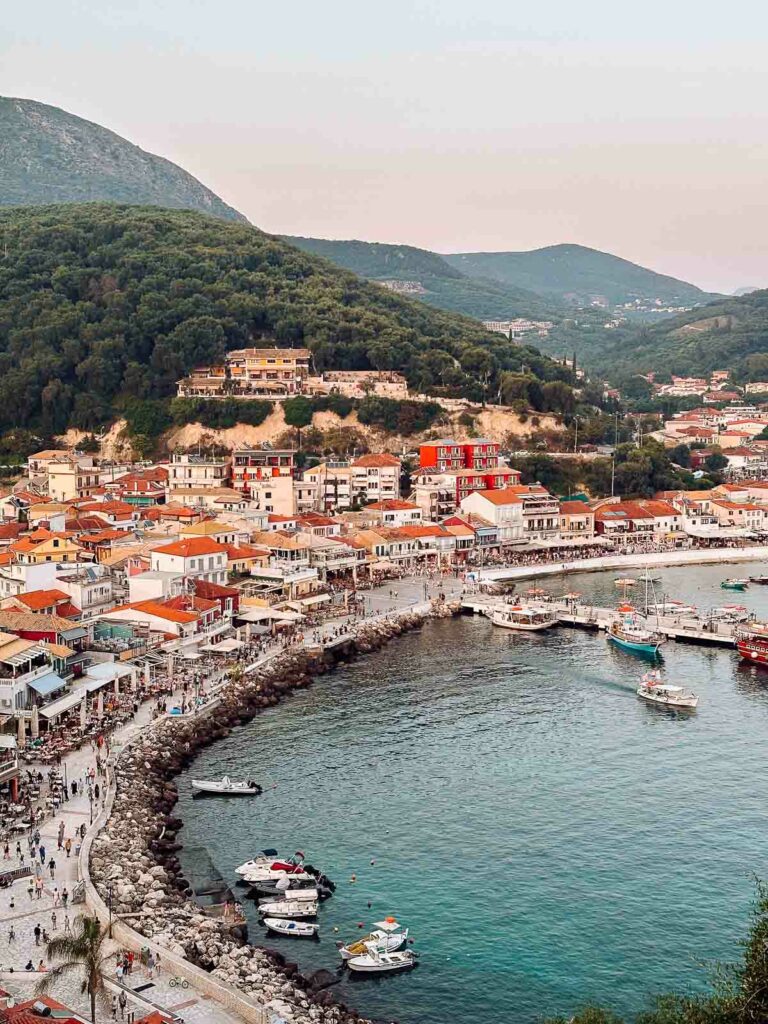 The quaint harbor of Parga, Greece, curves around a pebble beach, with terracotta rooftops and lush hillsides framing the serene Aegean Sea