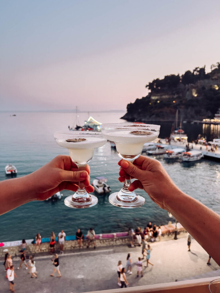 Cheers to the coastal vibes of Parga, Greece, with two margarita glasses clinking in a toast above a bustling seaside promenade at dusk