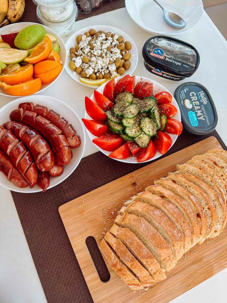 A spread of Greek breakfast on a boat, featuring fresh fruits, olives, cucumber-tomato salad, sliced bread, and traditional sausages, ready to be enjoyed on a MedSailors sailing tour in Greece