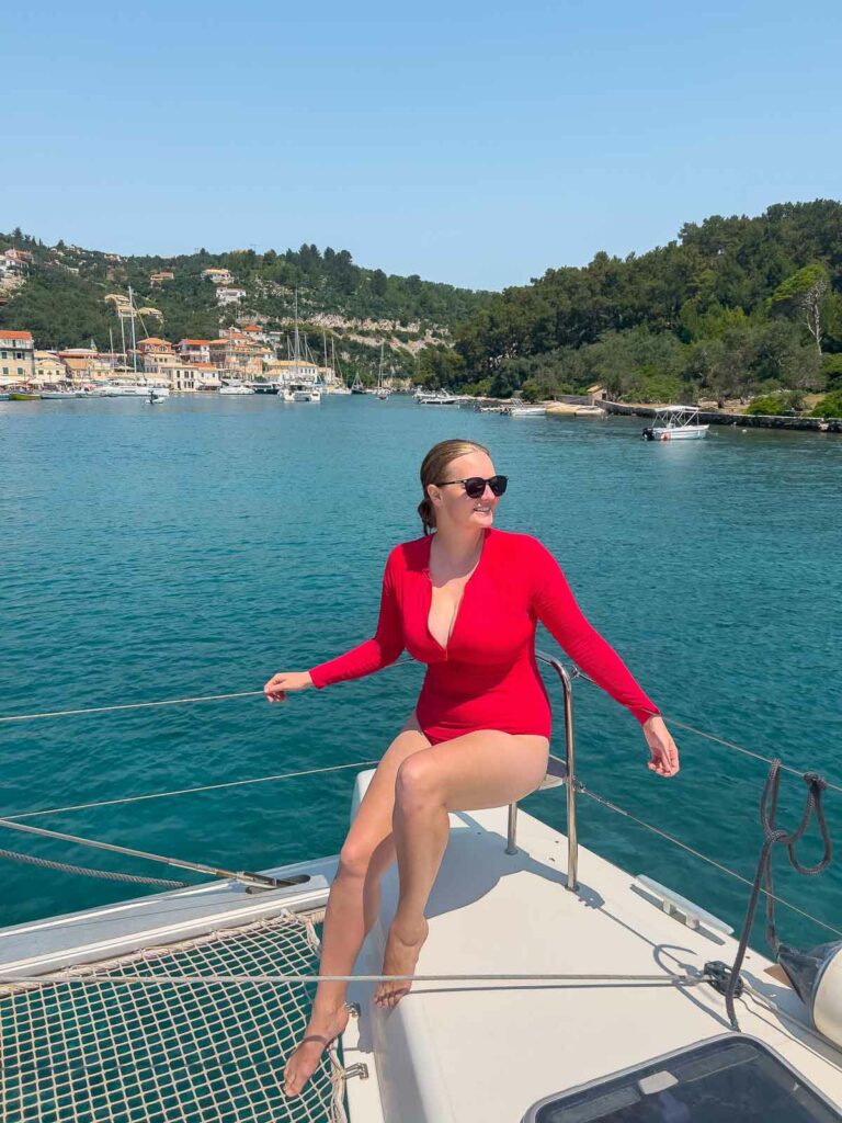 A person in a red swimsuit sits gracefully on the bow of a catamaran, with the picturesque coast of Greece and the azure Ionian Sea in the background