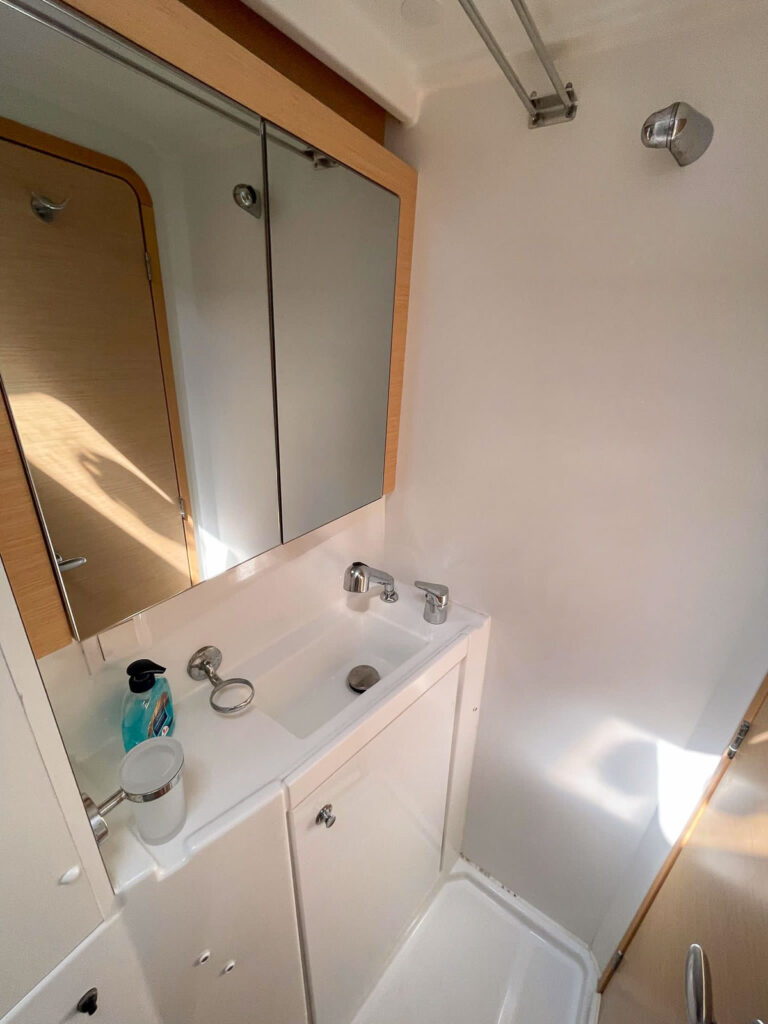 Compact and tidy bathroom on a MedSailors catamaran, featuring a white sink, mirror, and shower facilities, all efficiently designed for comfort at sea