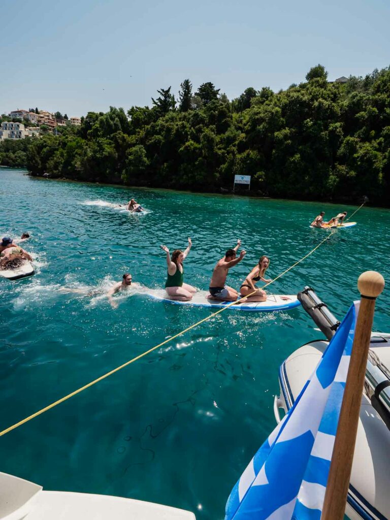 A group of people energetically participating in a paddleboard race, towed by a boat flying a Greek flag, with lush green coastline in the backdrop