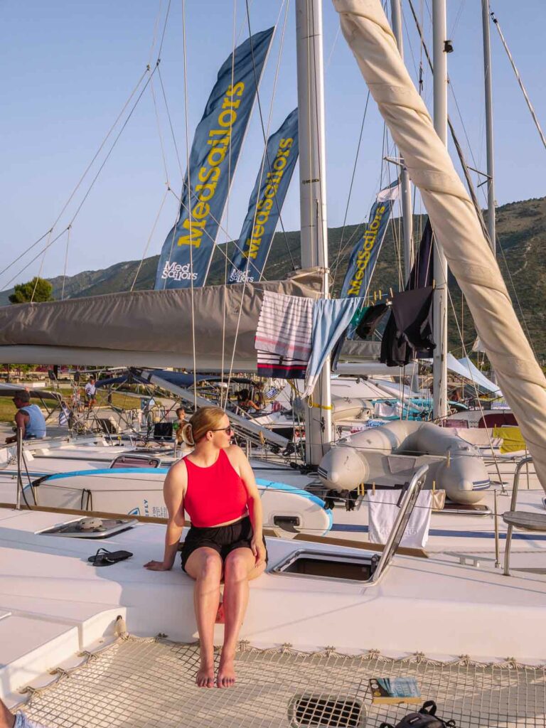 A traveler sits on the deck of a yacht, with MedSailors banners flying high above a fleet of yachts moored in a sunlit Greek marina