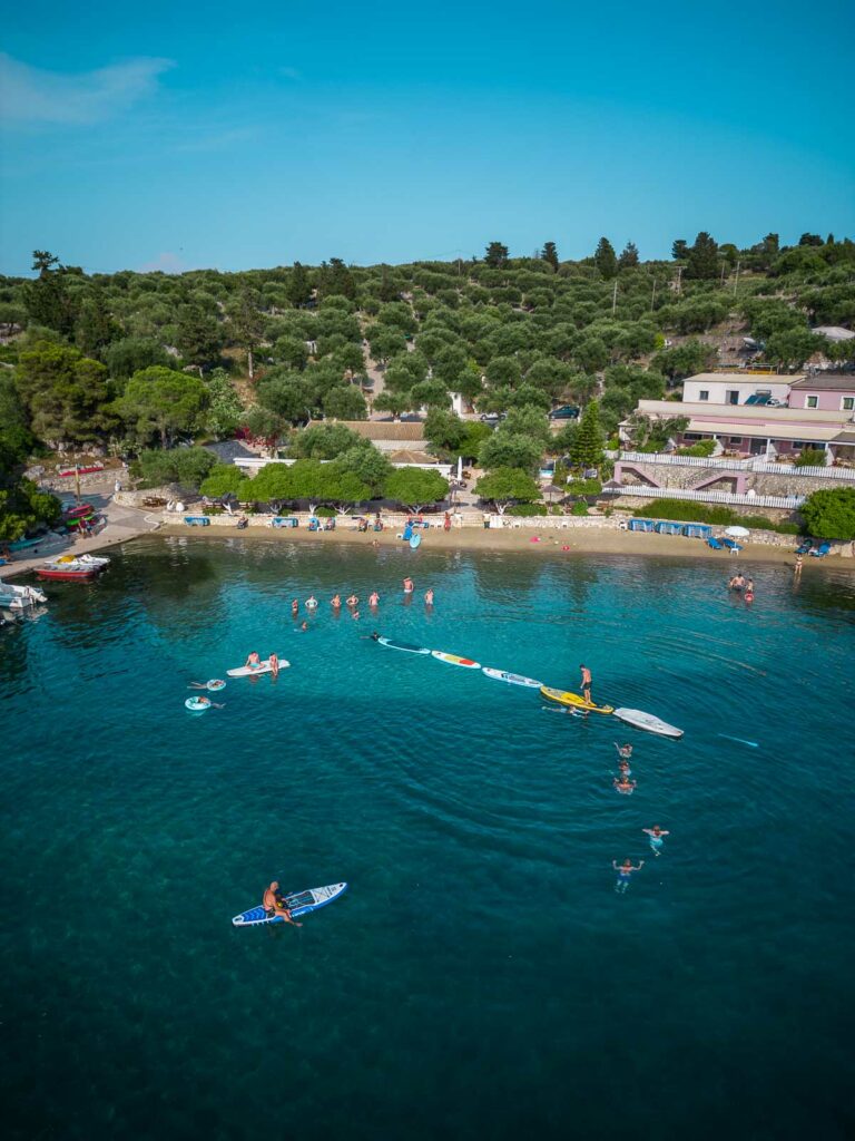 Aerial view of a paddleboard balancing competition with participants carefully navigating the calm blue waters off a verdant Greek island shore on a MedSailors tour