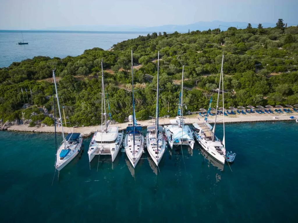 Aerial view of a fleet of Medsailors catamarans and yachts moored side by side along a lush green coastline on the clear blue waters of the Ionian Sea