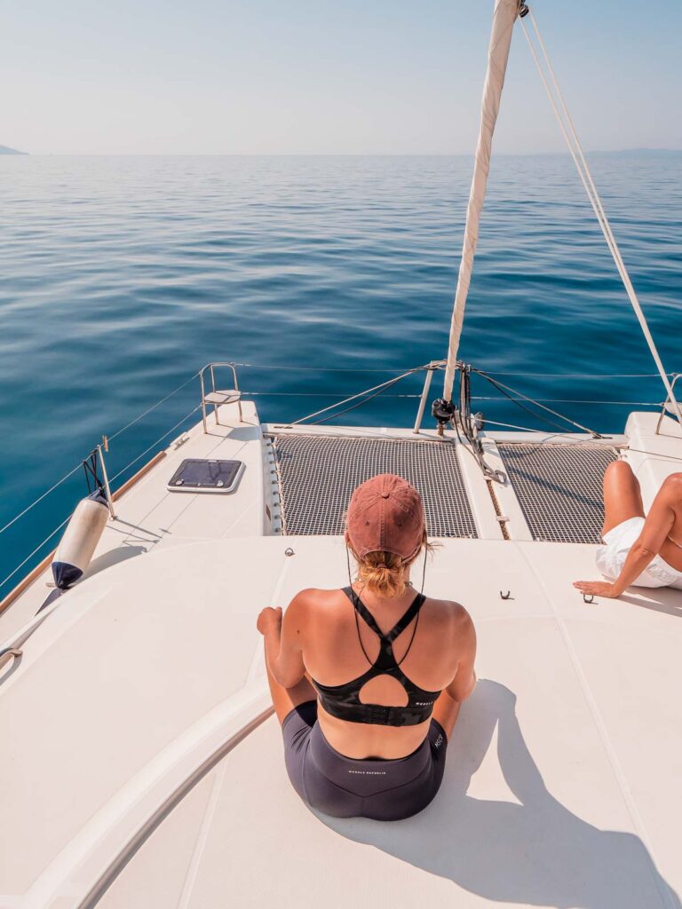 A woman sits peacefully on the deck of a catamaran, gazing out over the expansive blue waters, enjoying the calmness of a sailing adventure in Greece