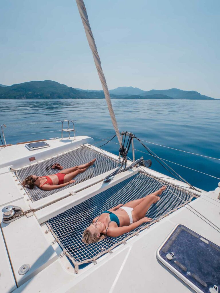 Two people relaxing on the net of a catamaran, basking in the sun against a backdrop of the calm blue waters of Greece during a MedSailors tour