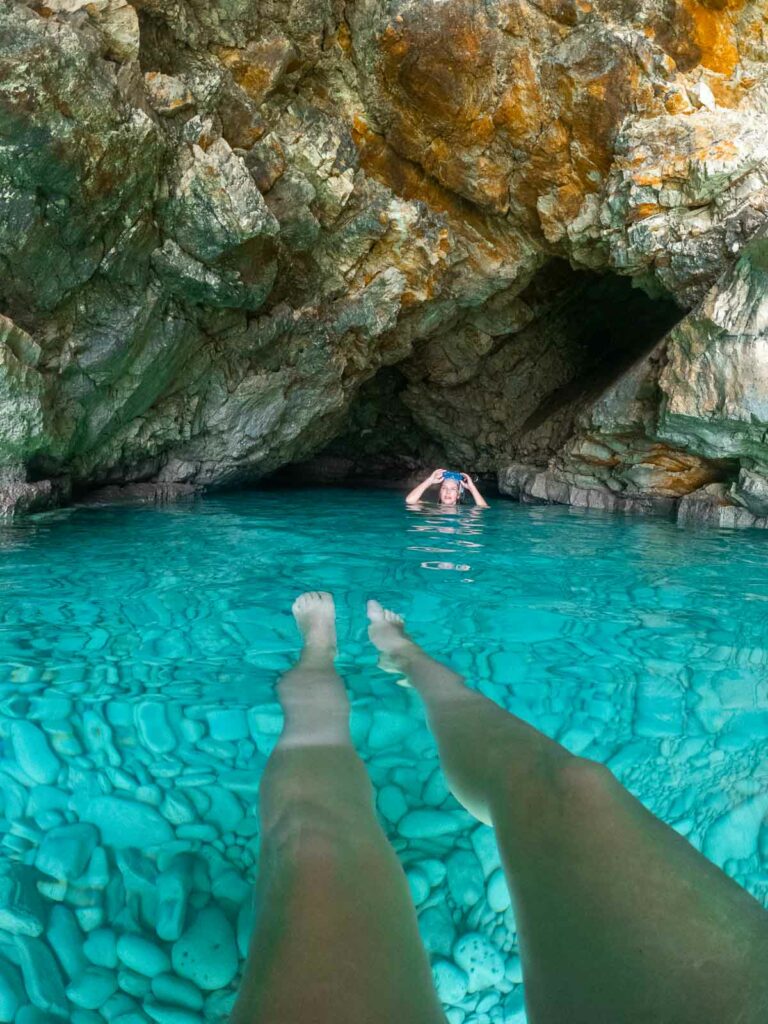 Crystal-clear turquoise waters in a Greek cove, viewed from above a swimmer's submerged legs, with a rocky cave entrance in the background