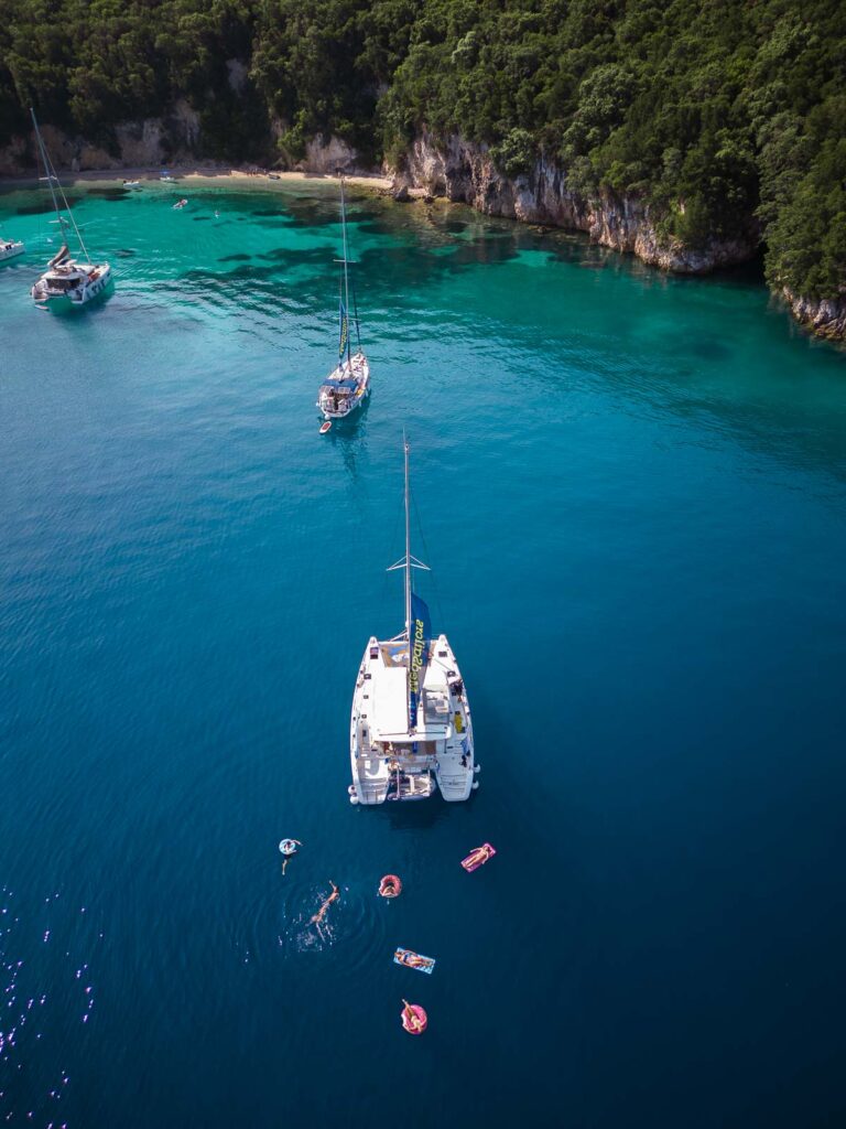 An aerial view of a MedSailors catamaran anchored in the clear blue waters of Greece, with swimmers and floaties around, epitomizing a Mediterranean summer