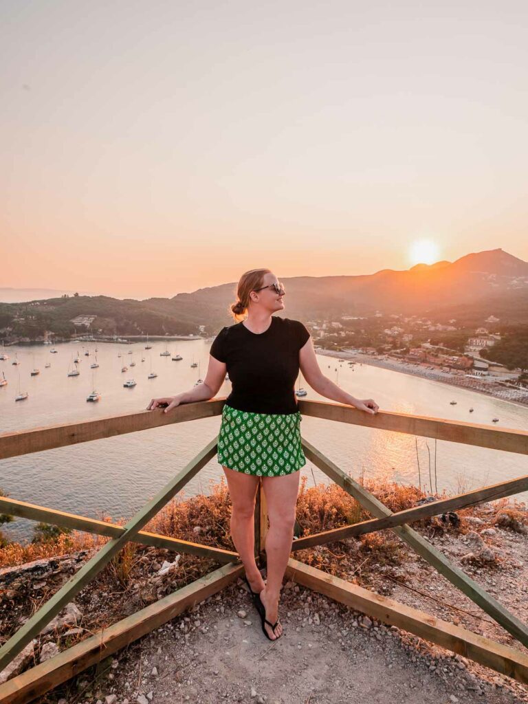A person stands at the top of the fort in Parga, Greece, taking in the stunning view of a sun setting over the harbor dotted with boats, the horizon painted with warm hues