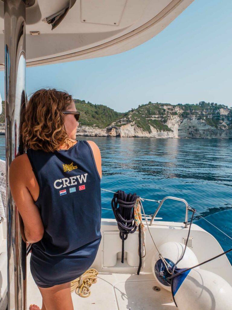 A member of the yacht crew, dressed in a 'CREW' tank top, leans thoughtfully against the rail, looking out over the tranquil blue waters of the Greek islands during a MedSailors sailing tour