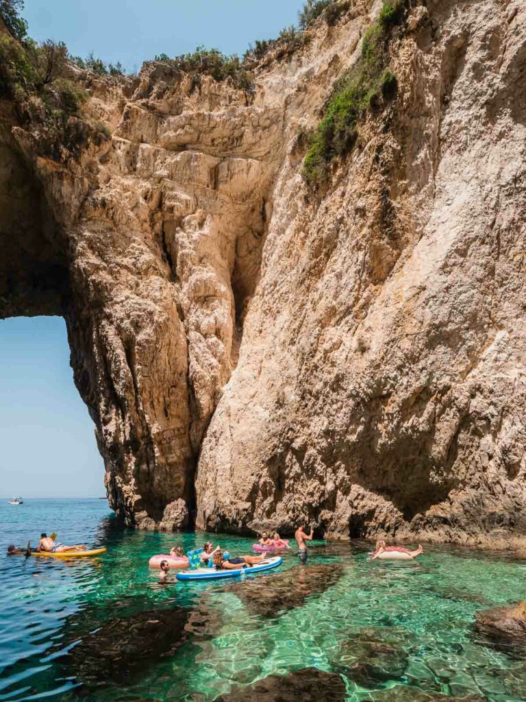 Visitors on colorful floaties enjoy the clear waters near a stunning natural rock arch on an island close to Corfu, Greece, with the rugged cliff face rising dramatically above them