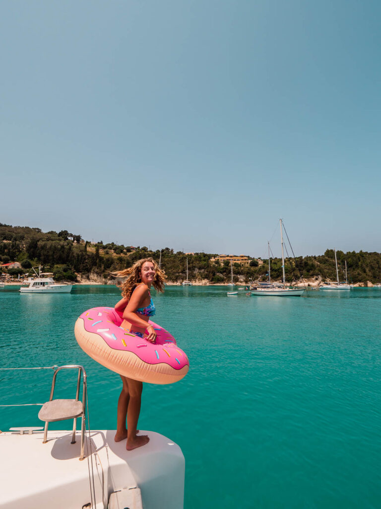 A joyful traveler stands on the edge of a catamaran holding a pink donut-shaped floatie, ready to enjoy the turquoise waters of a secluded Greek cove