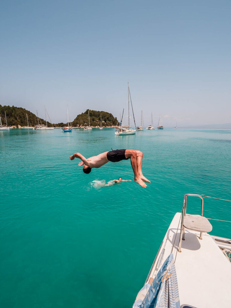 Person doing a backflip off the side of a MedSailors catamaran into the clear turquoise waters of the Ionian Sea, with other boats in the background