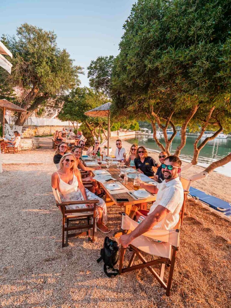 Travelers gathered around a rustic wooden table for dinner at a quaint Greek island restaurant, basking in the golden hour sunlight, with the serene beach and olive trees in the background
