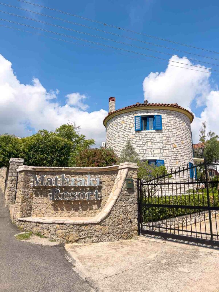 Entrance to Mathraki Resort in Corfu with a sign on a stone wall, featuring a traditional round stone tower with blue shutters against a blue sky with clouds