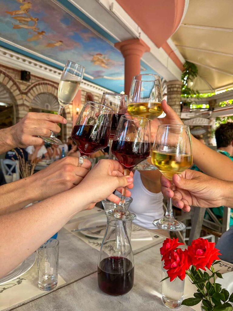 A festive toast with glasses of wine raised high among friends, with a decorative restaurant ceiling overhead and red roses on the table, encapsulating the joy of dining in Greece