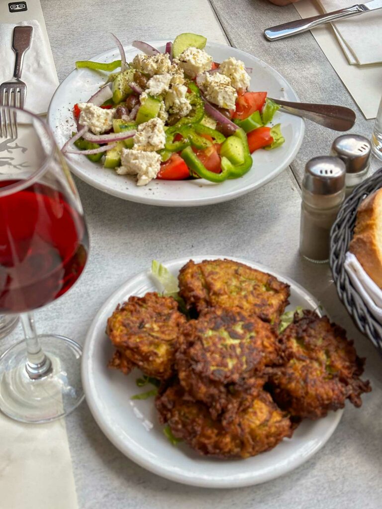 A traditional Greek meal laid out on a table, featuring a plate of crispy zucchini fritters, a fresh Greek salad, and a glass of red wine, inviting a taste of local cuisine