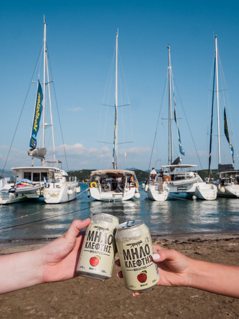 Two hands clinking cans of Minoan Ligo beer with a backdrop of MedSailors yachts anchored at a marina, celebrating the sailing lifestyle in Greece