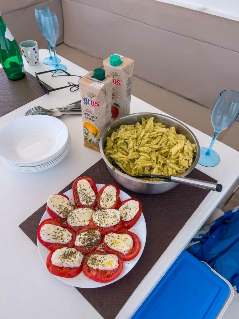 A yacht lunch spread featuring a bowl of pesto pasta and a plate of tomato and mozzarella bruschetta, with refreshing drinks ready on the side, during a MedSailors Greece tour