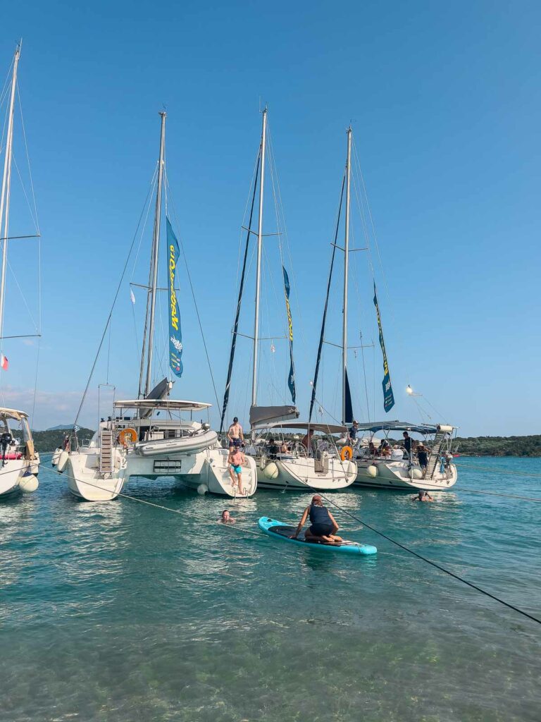 A line of MedSailors yachts moored in crystal-clear waters with people enjoying paddleboarding in the foreground, showcasing a vibrant aquatic activity in Greece