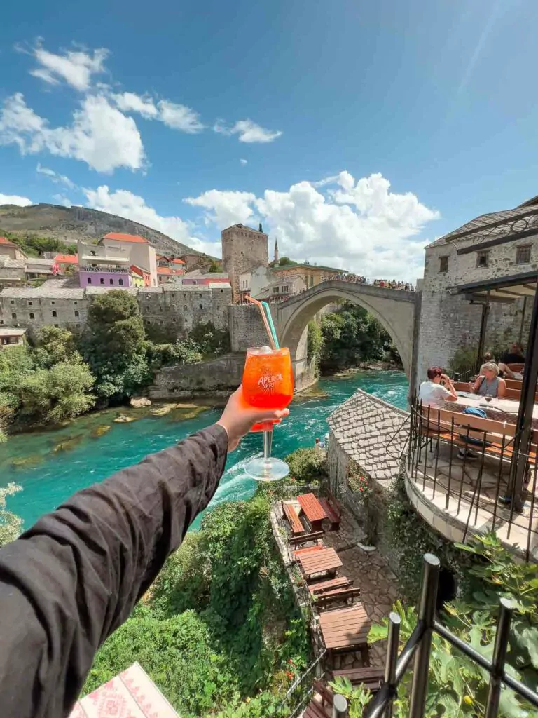 Person holding an Aperol Spritz with the iconic Stari Most bridge in the background, on a sunny day in Mostar, Bosnia