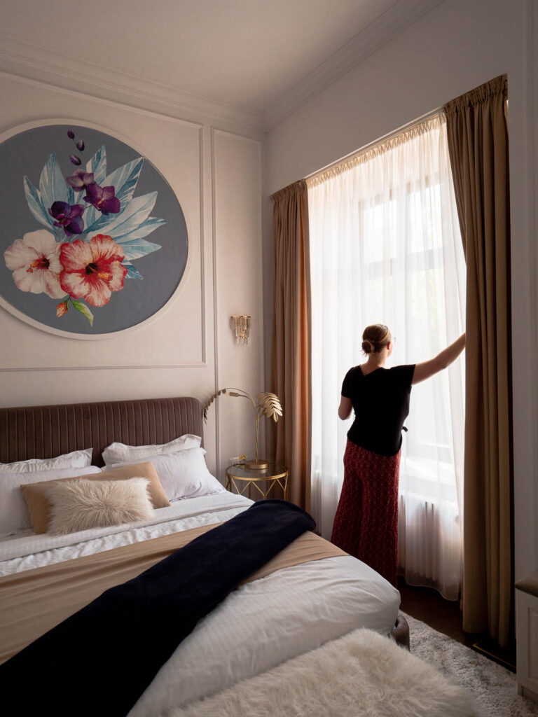 Elegant bedroom interior in Bucharest, Romania, with a large floral artwork above the bed, a woman opening sheer curtains to let light in, and tasteful decor featuring neutral tones and a touch of navy blue
