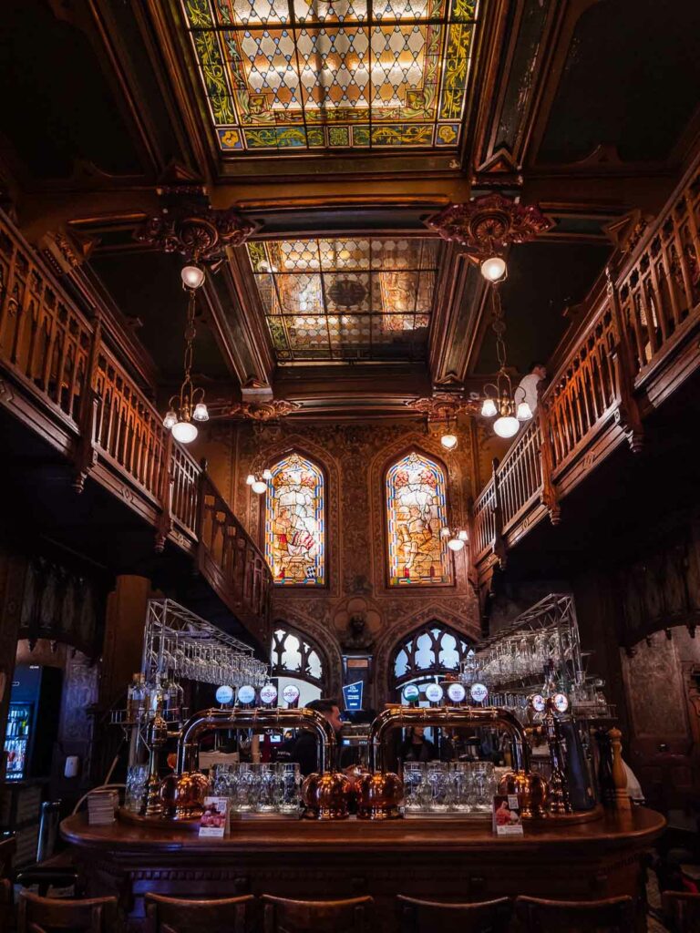 Interior of Caru' cu Bere in Bucharest, displaying its ornate woodwork and stained glass ceilings that illuminate the traditional bar setting with intricate details and historic charm