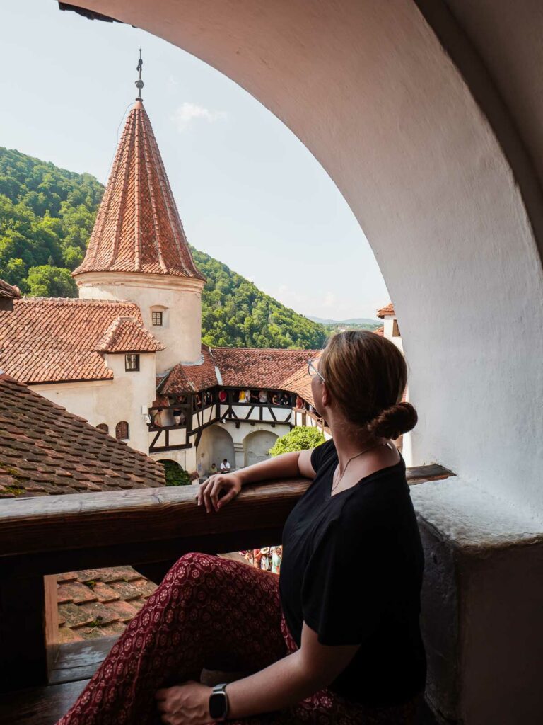 A woman gazing out from an arched window at Bran Castle in Transylvania, Romania, with a view of its distinctive turrets and the lush Carpathian Mountains in the background