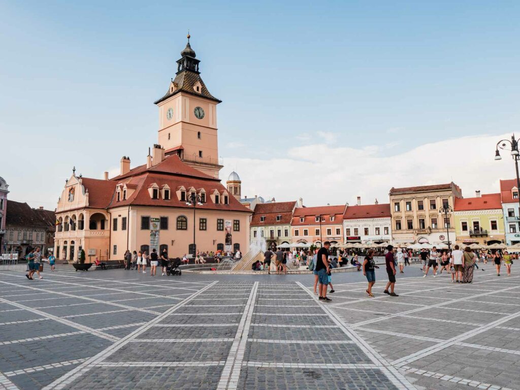 The bustling Council Square (Piața Sfatului) in Brașov, Transylvania, Romania, with the iconic Clock Tower of the History Museum, pastel-colored buildings, and visitors enjoying the vibrant atmosphere