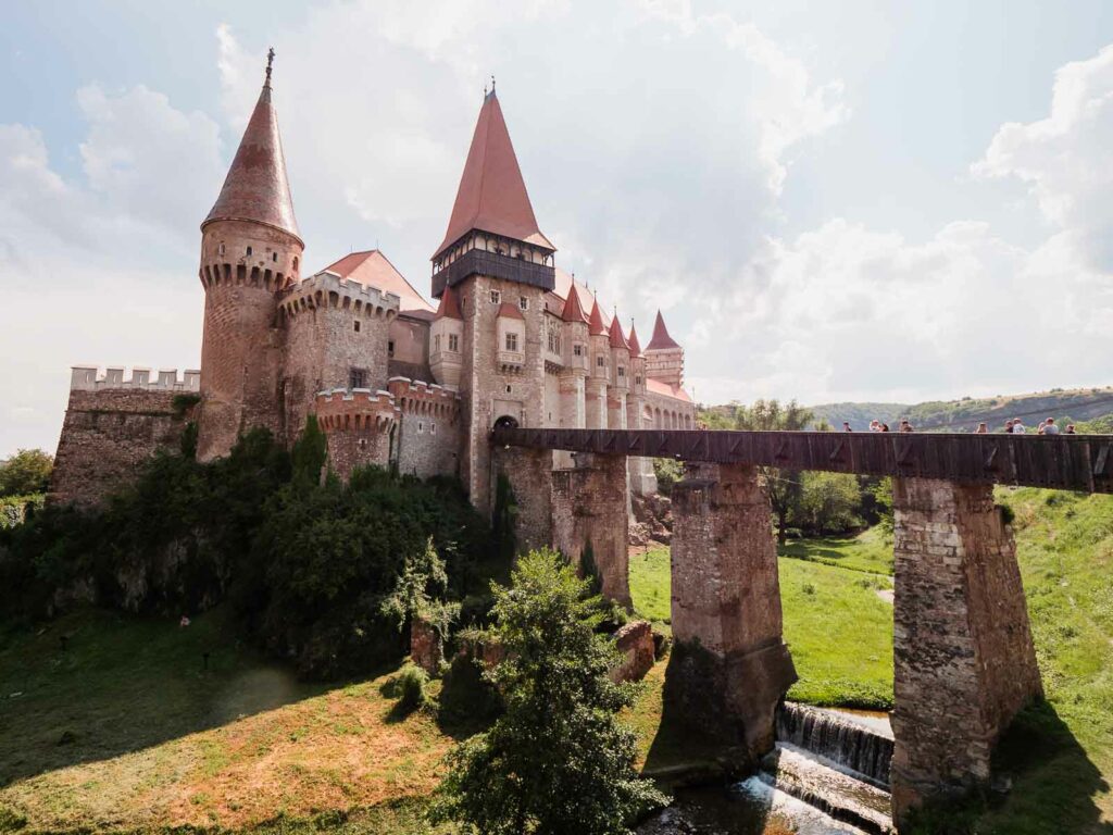 Corvin Castle, Romania, stands majestically against a blue sky, with its distinctive Gothic-Renaissance architecture, conical roofs, and a stone bridge over a small stream