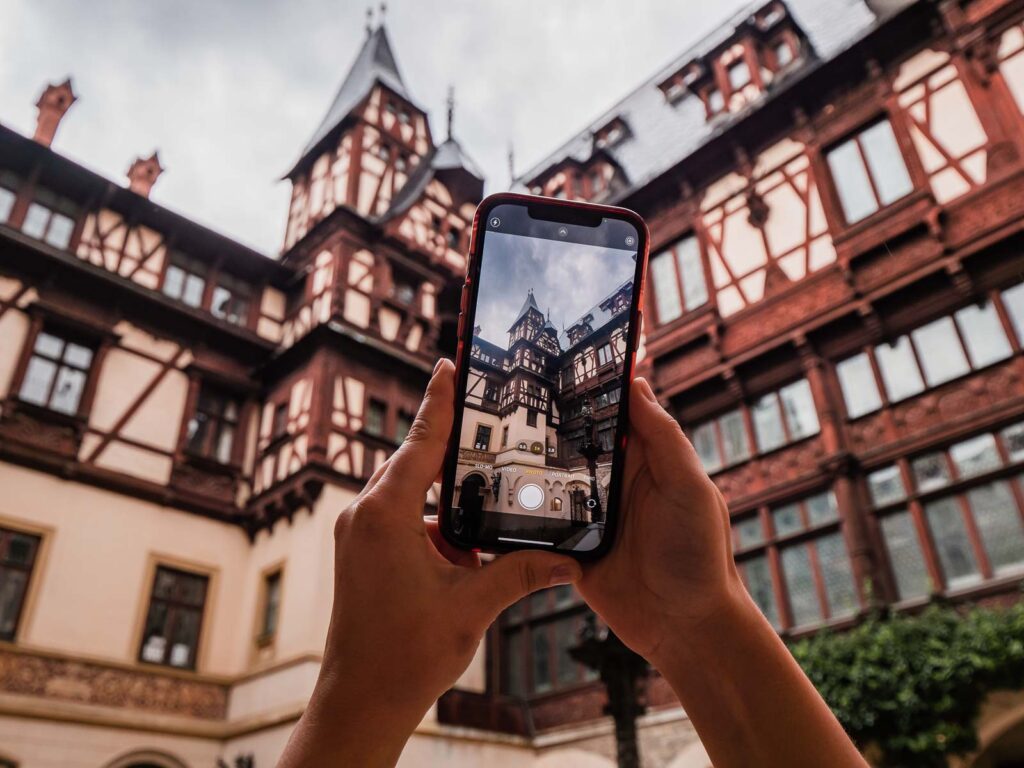Hands holding a smartphone capturing Peles Castle, Romania, through the screen, showcasing the castle's intricate architecture against a cloudy sky