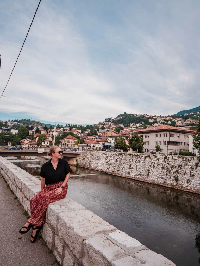 Woman sitting on a stone wall by the river in Sarajevo, Bosnia, with the cityscape and houses on the hill in the background, under a cloudy sky