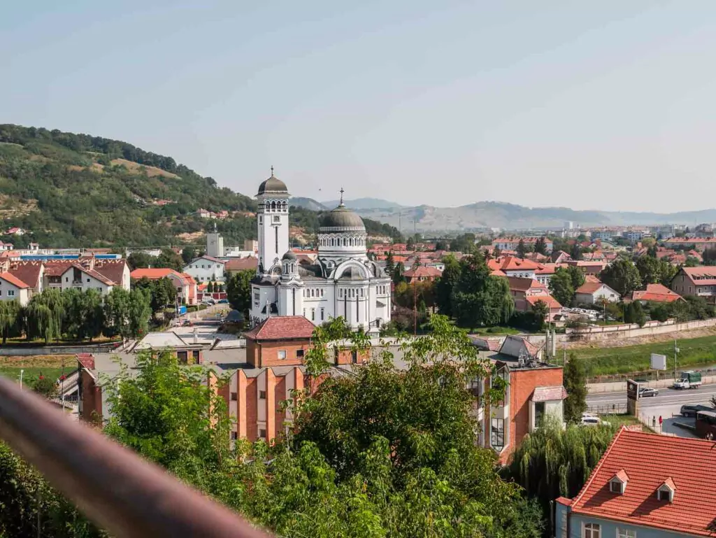Overlooking view of a town with traditional red-tiled rooftops and a prominent Orthodox church with gray domes and a bell tower, nestled among gentle hills
