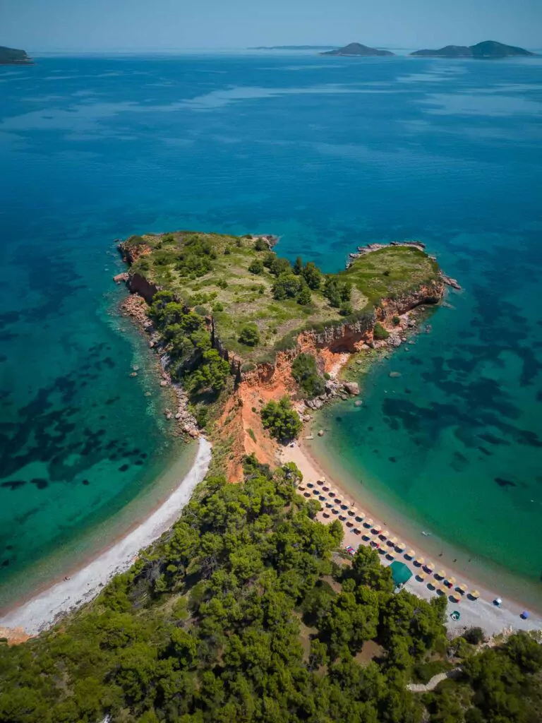 Aerial view of a Kokkinokastro Beach in Alonissos, showcasing the island's pristine travel appeal with crystal-clear turquoise waters, boats gently bobbing by the shore, and characteristic red-roofed buildings amid lush greenery. If you're planning a trip to Greece, this island has to be on your itinerary!