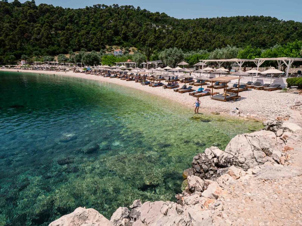 Scenic Leftos Gialos beach in Alonissos, with a curving shoreline of clear turquoise waters and white pebbles, complemented by rows of sunbeds under natural wood umbrellas, offering a peaceful beachside escape