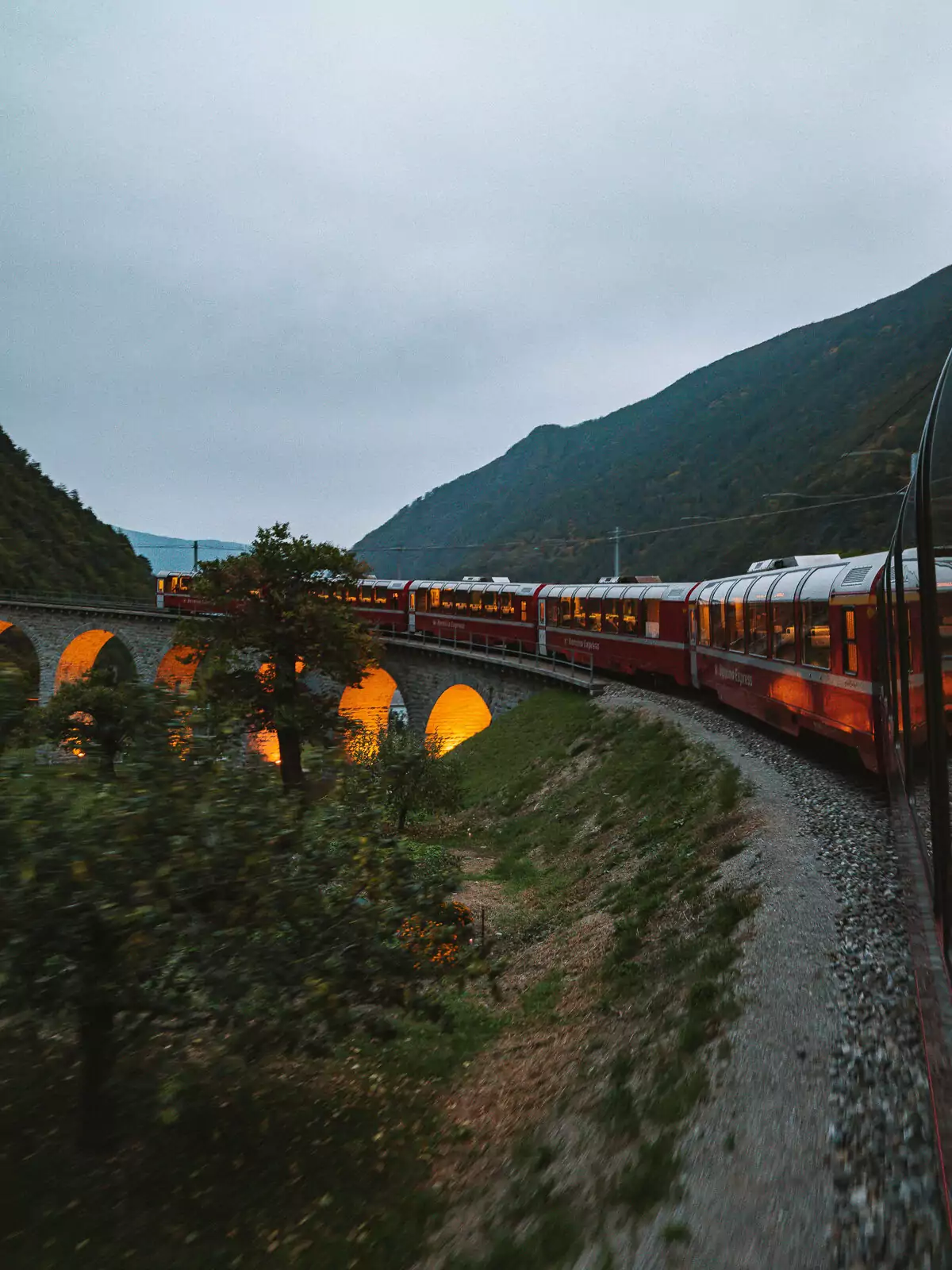Bernina Express train lights glowing warmly as it travels through mountain valley at dusk