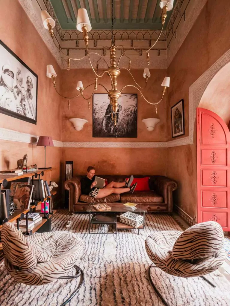 An elegantly decorated lounge in the Indian Palace Riad, Marrakech, with plush seating, patterned rugs, and striking chandeliers, offering a blend of Moroccan and Indian decor that invites relaxation and conversation