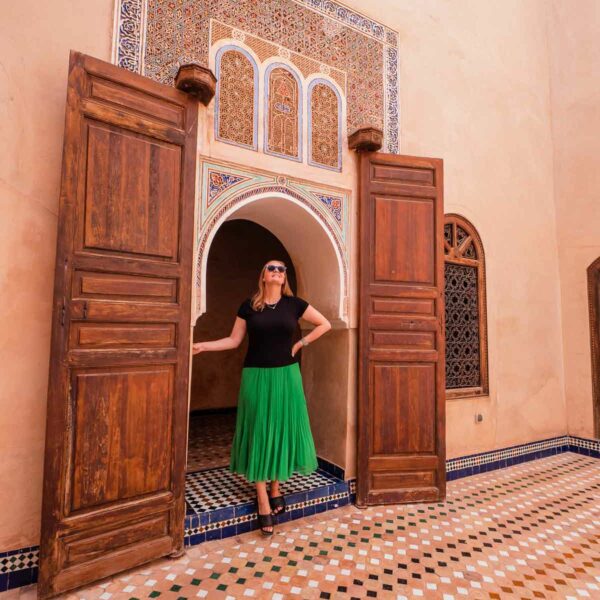 A visitor stands at the entrance of an ornately decorated doorway at Bahia Palace in Morocco, with intricate mosaic tiles and richly carved wooden doors, highlighting the architectural grandeur of the palace