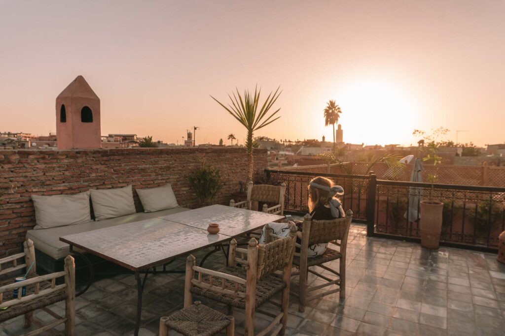 A tranquil sunset view from the rooftop of Riad Nirvana in Marrakech, with silhouettes of palm trees and minarets against the glowing sky, an idyllic setting to unwind after exploring