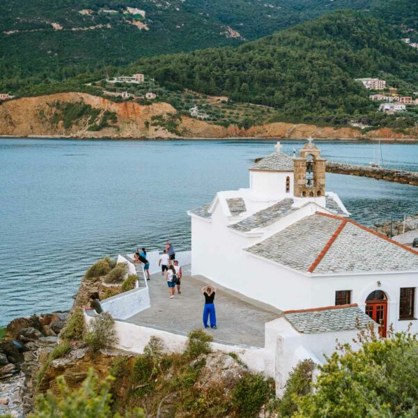 Tourists gather around the iconic main church of Skopelos, with its stark white walls and traditional architecture standing out against the backdrop of the island's lush hills and calm sea, a focal point in any Skopelos itinerary