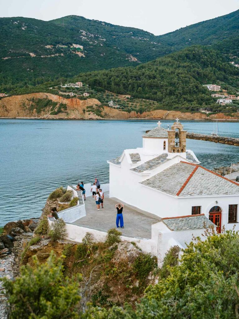 Tourists gather around the iconic main church of Skopelos, with its stark white walls and traditional architecture standing out against the backdrop of the island's lush hills and calm sea, a focal point in any Skopelos itinerary