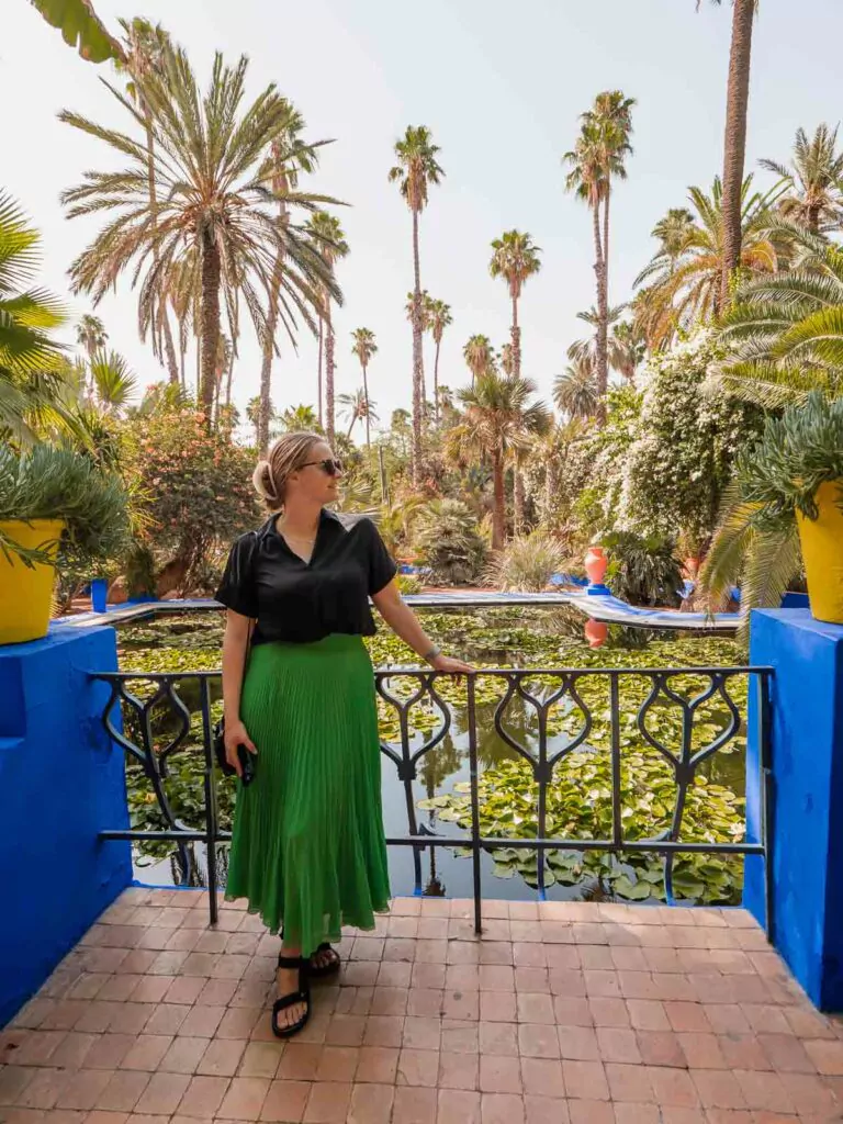 A solo traveller in a vibrant green skirt leans on a blue balcony railing at Jardin Majorelle, surrounded by lush palms, encapsulating the exploration of two days in Marrakech