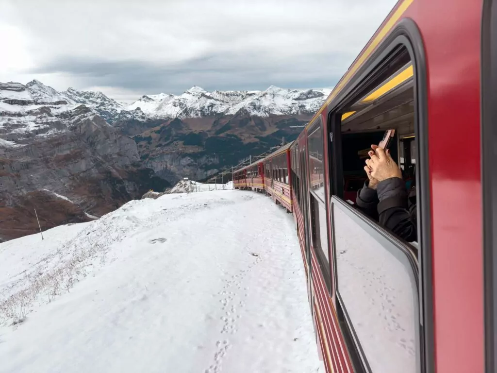 A passenger's hand reaches out of the window of the red train from Kleine Scheidegg to Jungfraujoch, capturing the essence of adventure amidst the snowy landscape and Alpine peaks