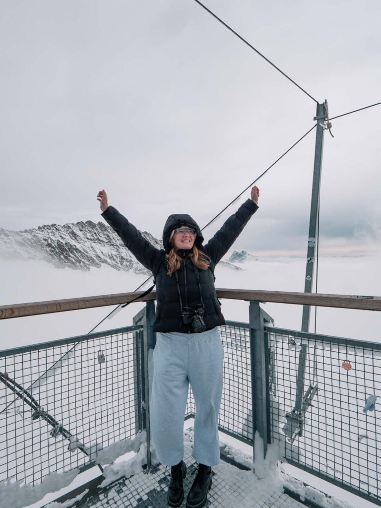 A jubilant visitor with arms raised stands on a viewing platform at Jungfraujoch, surrounded by the vast snow-covered peaks of the Swiss Alps, a memorable highlight on any Switzerland itinerary